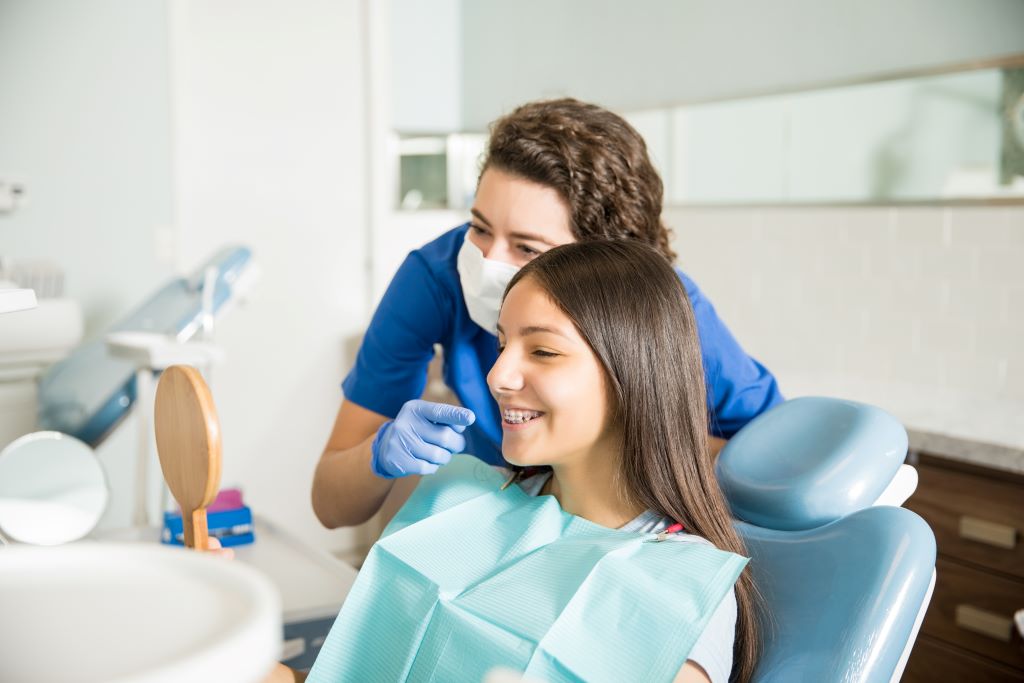 Na imagem, há uma jovem adulta, com os cabelos lisos, de aparelho dental, sorrindo e sentada na cadeira do dentista, atrás dela há uma dentista apontando para seu sorriso e explicando algo, ambas olham para um espelho que a jovem está segurando.