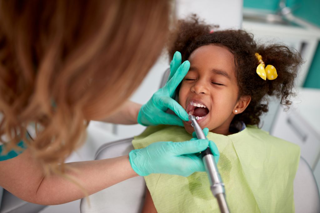 Na imagem, uma criança com maria chiquinhas, sentada na cadeira do dentista e a dentista está com o aparelho em seus dentes.